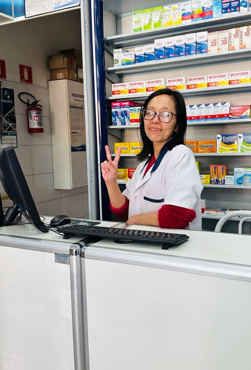 Farmacêutica sorrindo no balcão da Celeste Drogaria Popular, com as prateleiras de medicamentos ao fundo